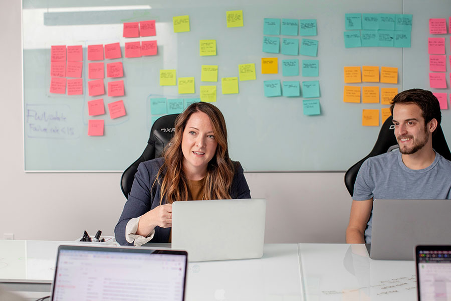 two people sitting in a conference room at laptops with sticky notes on a whiteboard behind them