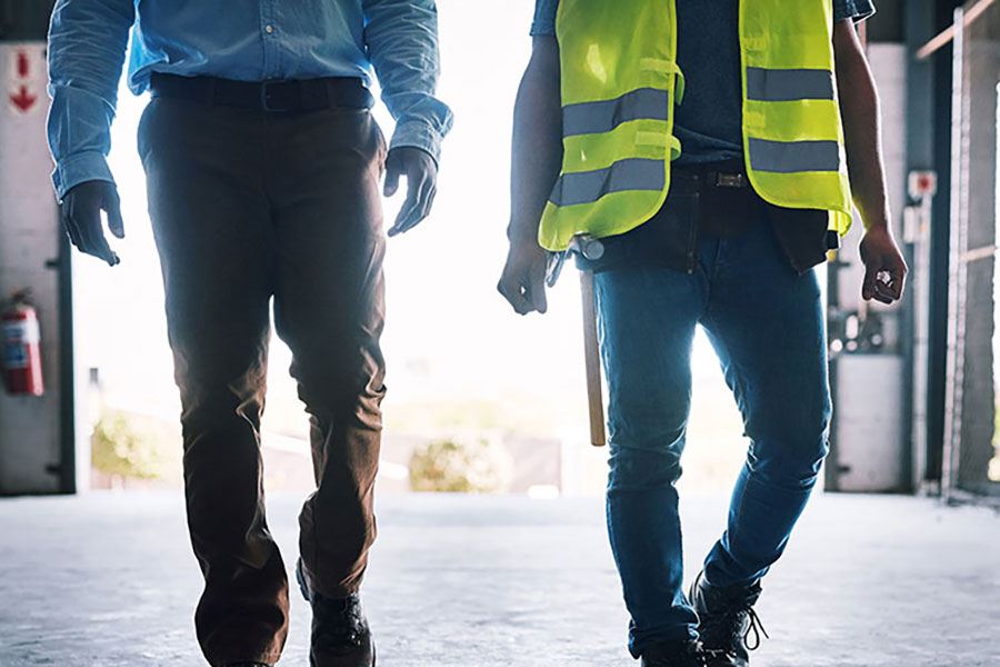 lower half of two people walking in a concrete setting one wearing high visibility and one wearing a suit