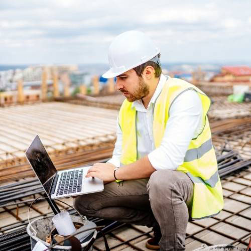 Man in yellow construction vest on site and typing on a laptop